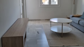 A modern living room with a wooden bench, a round coffee table, a sofa, a patterned rug, a hanging lamp, and a window with a view of a building with a tiled roof. - Apartment 5 minutes walk from the Fair. Ideal for couples. (Jerez de la Frontera)