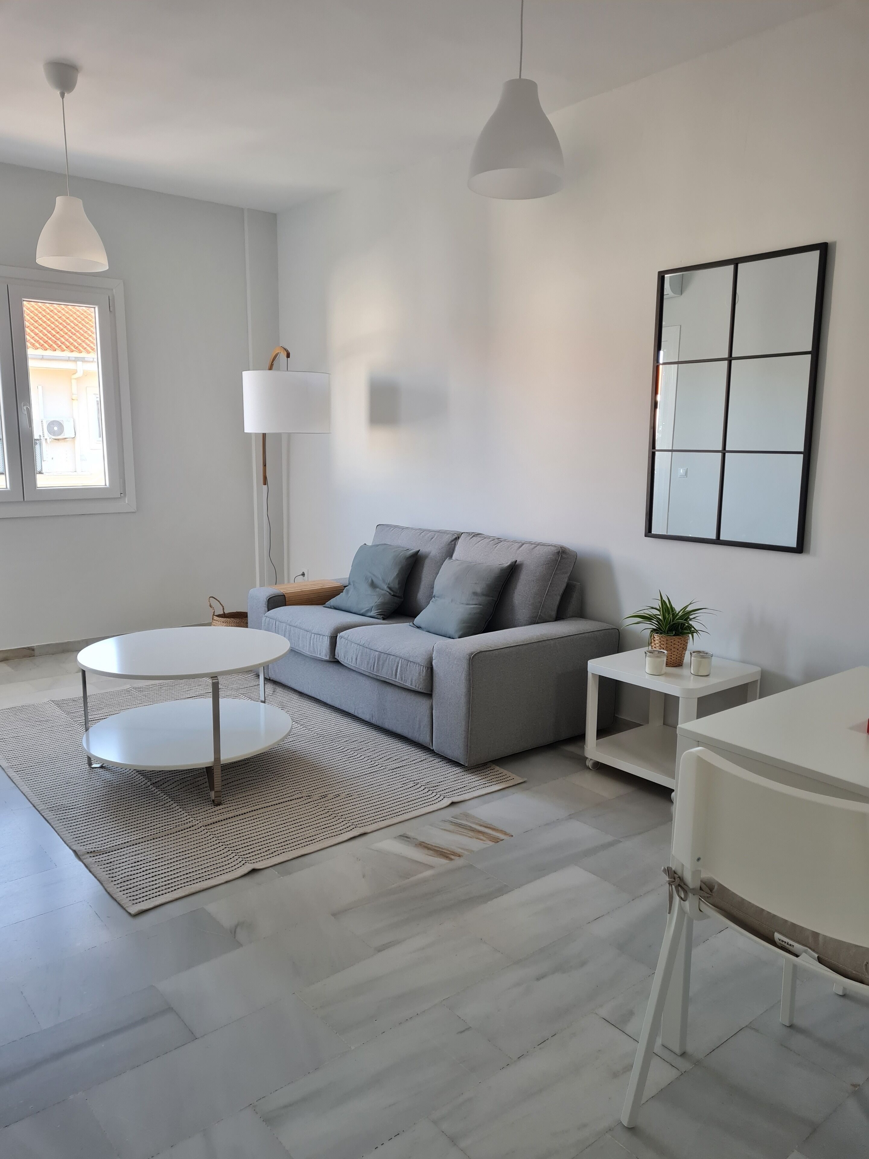Un salon moderne avec un canapé gris, une table basse ronde blanche, une table d’appoint avec une plante et un grand miroir au mur.