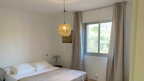 Et soveværelse med seng, sengetæppe, et vindue med gardiner og en lampe på væggen.