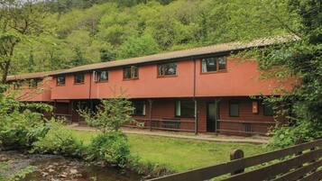 Un edificio de dos pisos con fachada roja, rodeado de exuberante vegetación y un pequeño arroyo.