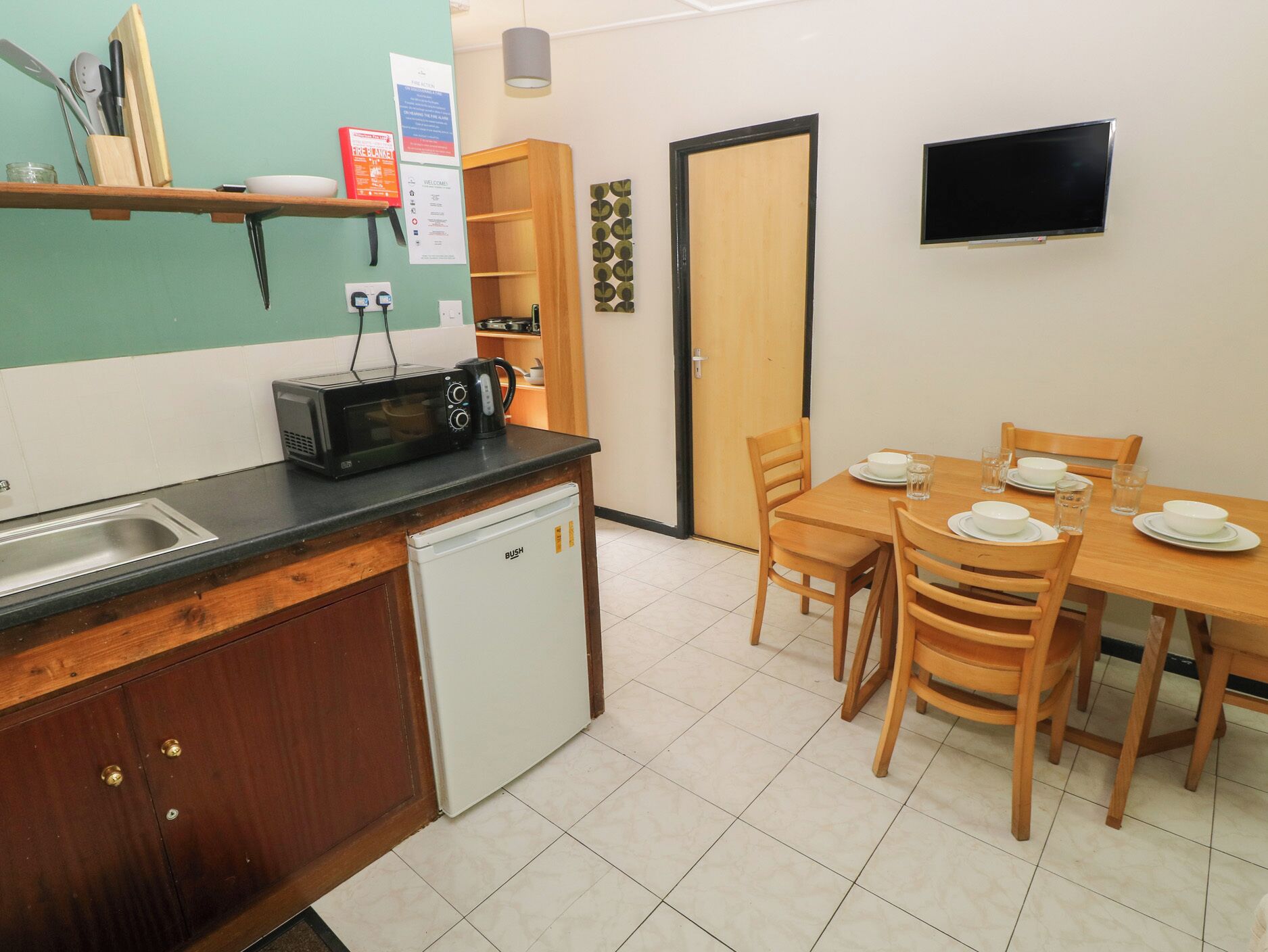 A compact kitchen with a microwave, sink, and a small refrigerator. There is a dining table with chairs and a television mounted on the wall.