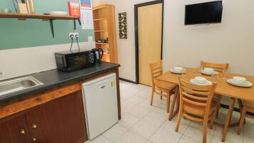 A compact kitchen with a microwave, sink, and a small refrigerator. There is a dining table with chairs and a television mounted on the wall.