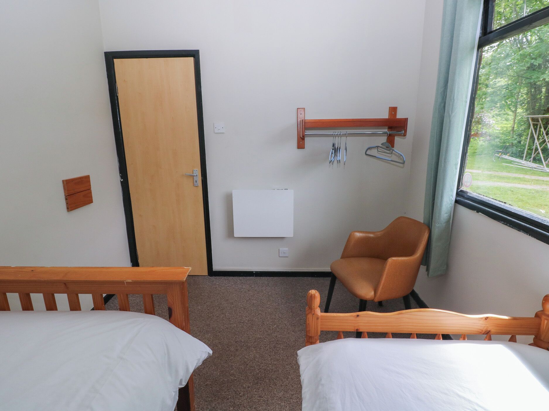 A room with two wooden beds, a chair, a door, a wall-mounted heater, and a window with a view of greenery.
