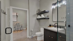 Ein modernes Badezimmer mit Toilette, Waschbecken und Duschkabine aus Glas. Über der Toilette befinden sich Regale und im angrenzenden Raum ein runder Tisch mit Stühlen.