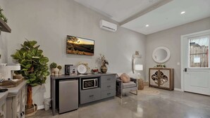A modern living room with a grey sofa, a small cabinet, a dining table with chairs, a large potted plant, and a wall-mounted air conditioner. - Poplar Place 3 - Hot Tub & Close to Downtown (Fredericksburg)
