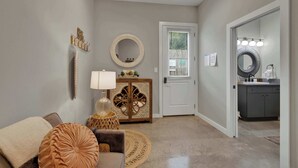 A living room with a sofa, a round rug, a lamp, a decorative mirror, and a door leading to a bathroom with a vanity and mirror. - Poplar Place 3 - Hot Tub & Close to Downtown (Fredericksburg)
