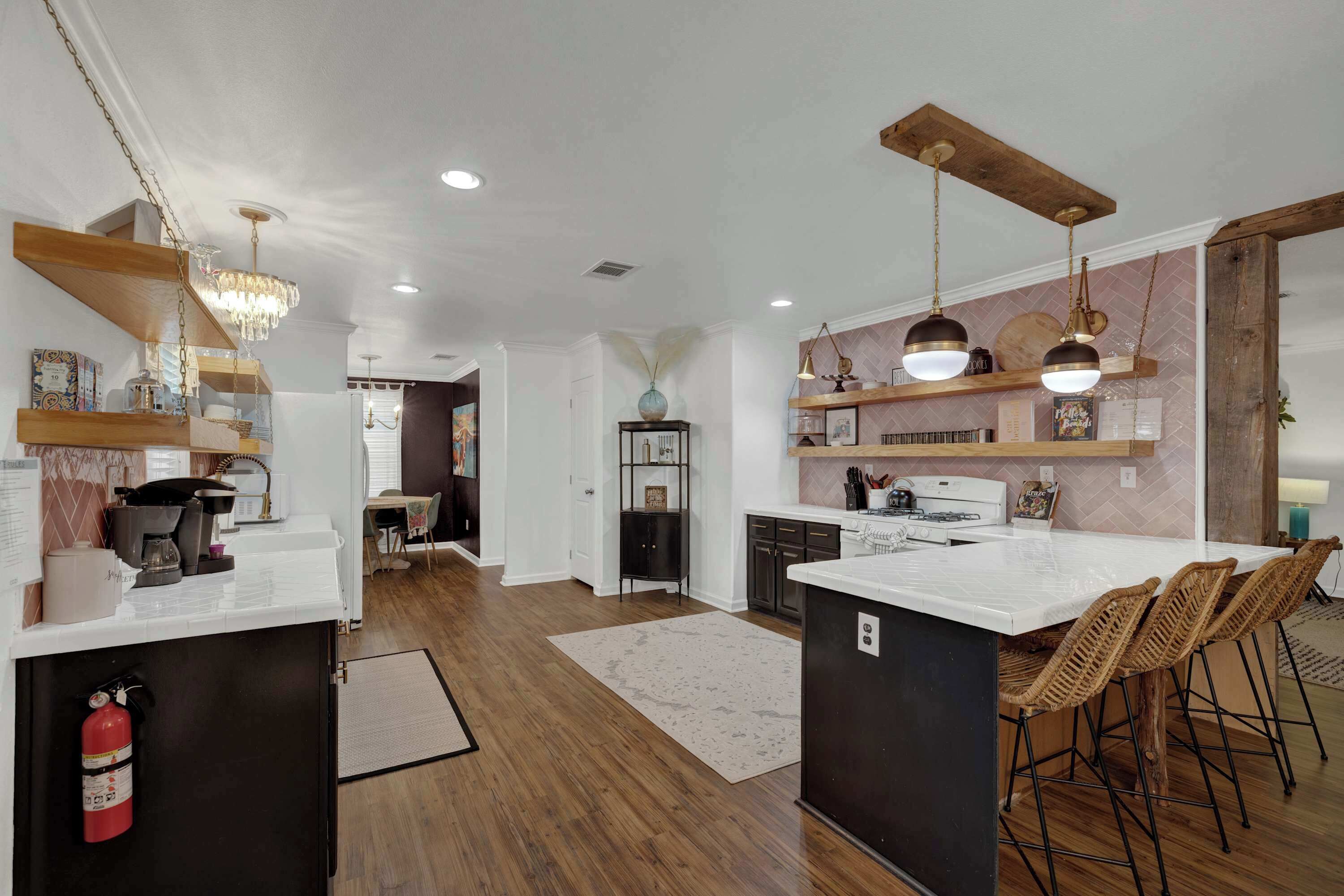 A modern kitchen with wooden flooring, white countertops, and bar stools. There is a dining area with a table and chairs, and a kitchen island with a sink.