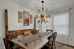 A dining area with a wooden table, chairs, a woven placemat, and a modern chandelier. - Poplar Place Mod - Hot tub & Fire Pit (Fredericksburg)