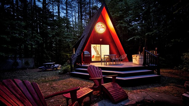 Eine dreieckige Holzhütte mit einer Terrasse, Sitzgelegenheiten im Freien und einer Feuerstelle.