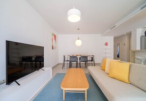 A modern living room with a flat-screen TV, a sofa with yellow cushions, a wooden coffee table, and a dining area with a table and chairs. - Arenal Dream Glow Apartment Jávea (Javea / Xàbia)