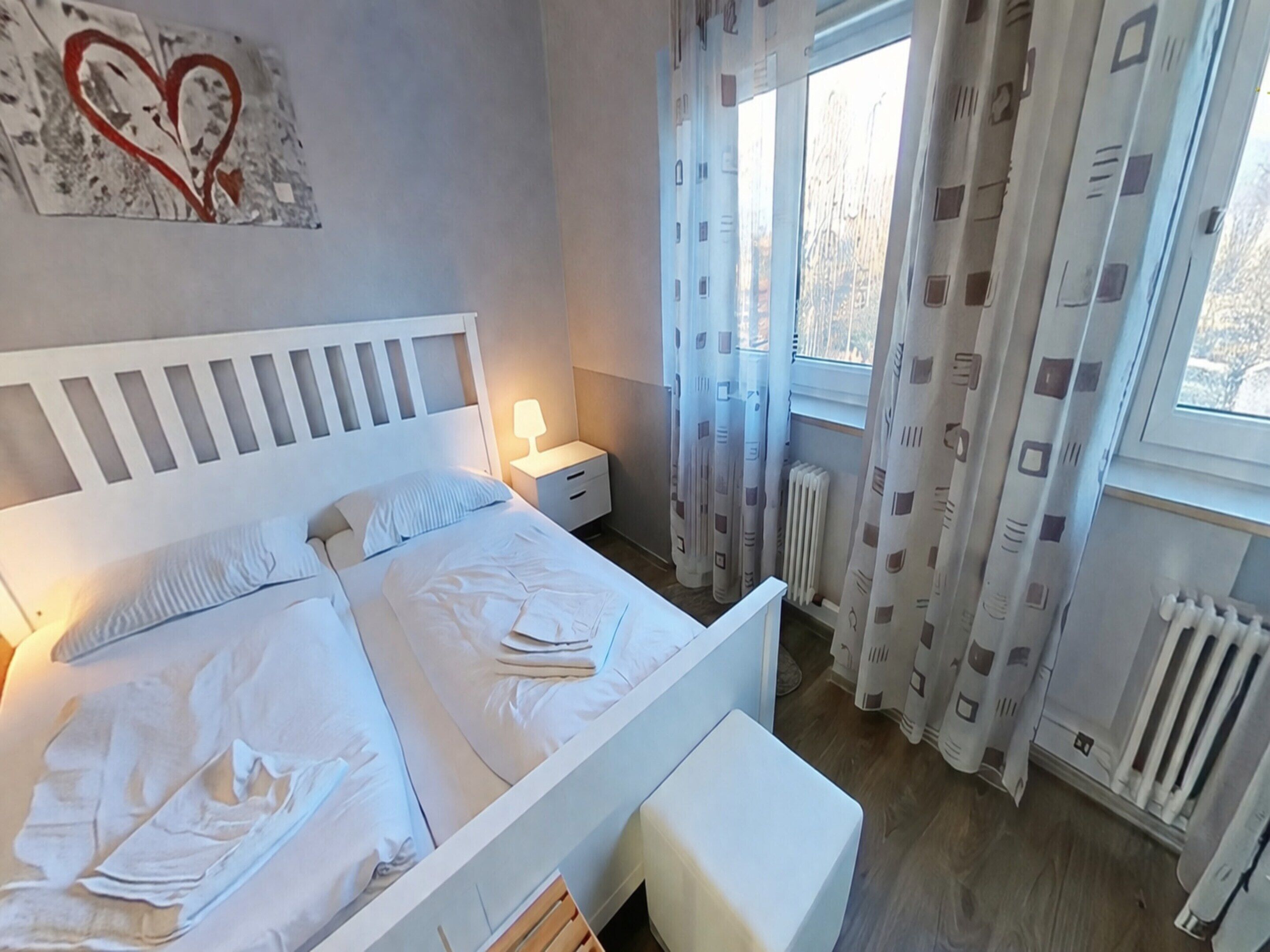 Quarto com cama, mesa de cabeceira e janela com cortinas.