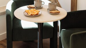 Une table ronde avec une tasse et une soucoupe, une assiette avec des tranches d’orange et une tranche de pain. Il y a deux chaises vertes autour de la table.