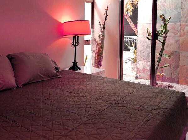 Une chambre à coucher avec un lit, des oreillers, une table de chevet, une lampe et une vue sur un cactus, visible à travers une porte vitrée.