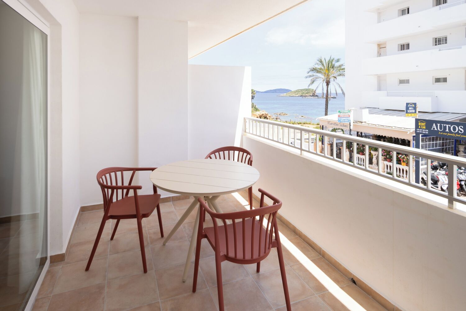Un balcón con una mesa redonda y cuatro sillas, con vistas a una zona costera y el mar, y un edificio con un letrero que dice 'RENT A CAR' y 'AUTOS'.