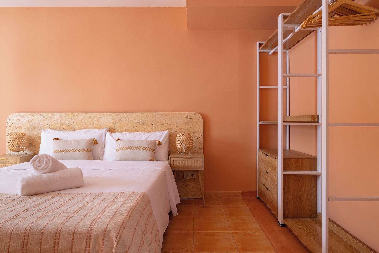 Een slaapkamer met een bed, kussens, een nachtkastje, een lamp en een houten garderobe.