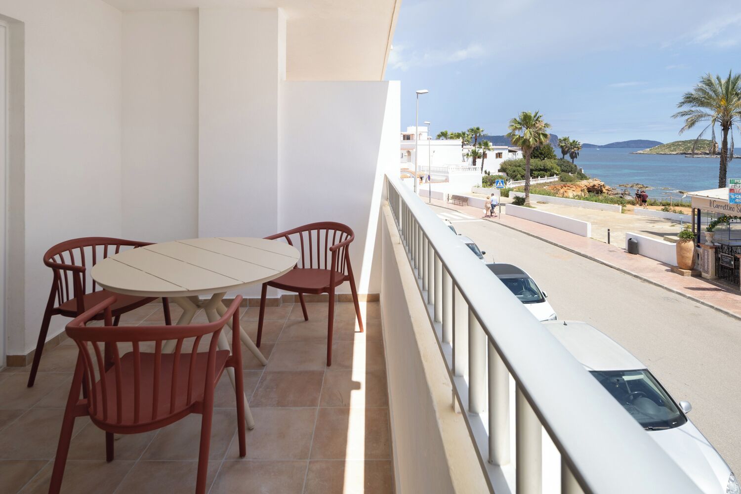 A balcony with a table and chairs, overlooking a coastal road with cars and palm trees.