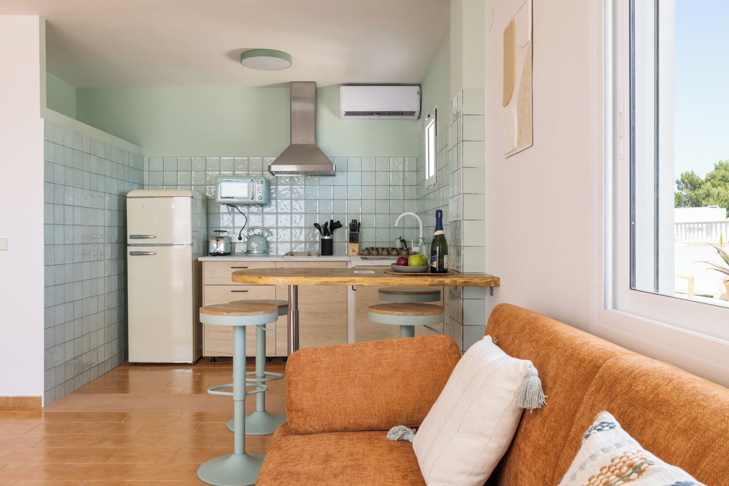 Una cocina moderna con refrigerador, estufa y lavamanos, un taburete de madera y vista al exterior a través de una ventana.