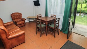 Una habitación con un sofá de cuero marrón, una mesa de comedor de madera con sillas, un televisor pequeño montado en la pared y un ventanal con cortinas verdes.