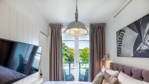 Quarto com cama, televisão, luz no teto, janela com cortinas e uma obra de arte na parede.