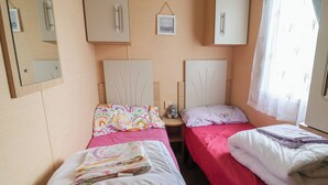 Ein Zimmer mit zwei Betten, jeweils mit unterschiedlicher Bettdecke, einem kleinen Nachttisch mit Teddybär, einem Wandspiegel und einem Fenster mit durchsichtigen Vorhängen.