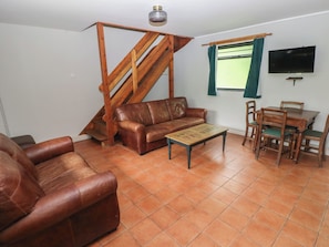 Una sala de estar con un sofá de cuero marrón, una mesa de centro de madera, una mesa de comedor con sillas, una escalera de madera y un televisor montado en la pared.
