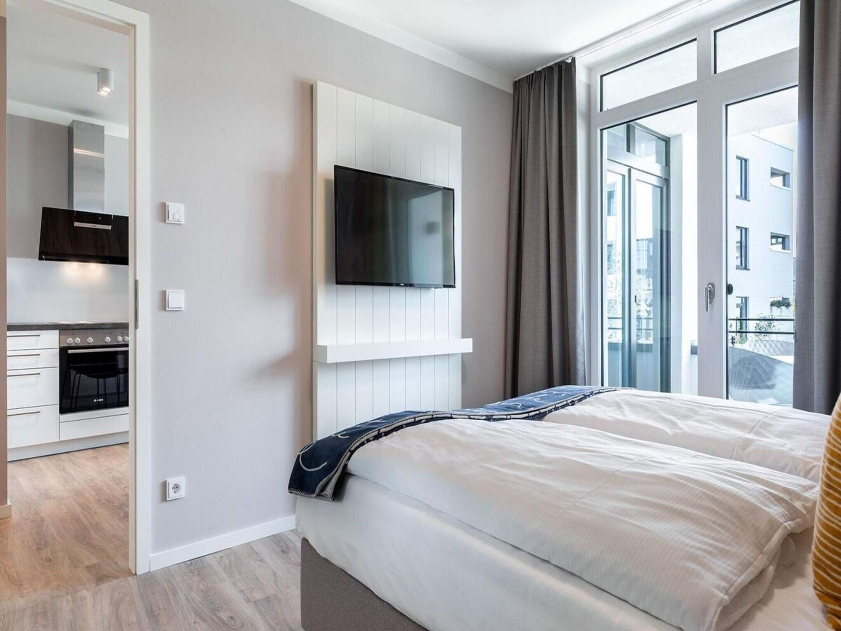 A modern hotel room with a bed, a TV mounted on the wall, a kitchen area, and large windows.