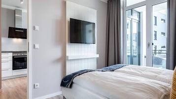 A modern hotel room with a bed, a TV mounted on the wall, a kitchen area, and large windows.