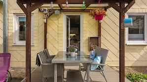 Eine überdachte Terrasse mit einem Glastisch, Korbstühlen und hängenden Blumenampeln.