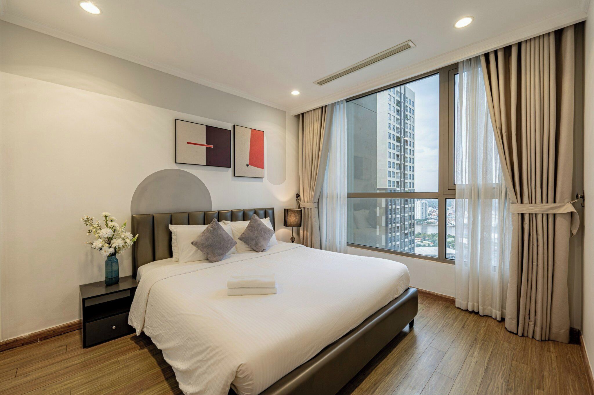 Une chambre d’hôtel moderne avec un grand lit, une table de chevet, un vase de fleurs et une vue sur le paysage urbain, visible à travers une grande fenêtre.
