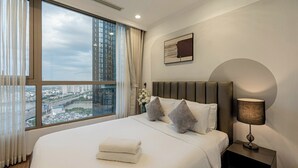 Quarto de hotel com uma cama grande, mesa de cabeceira com luminária, vista para a cidade através de uma janela ampla e uma moldura com imagem na parede.