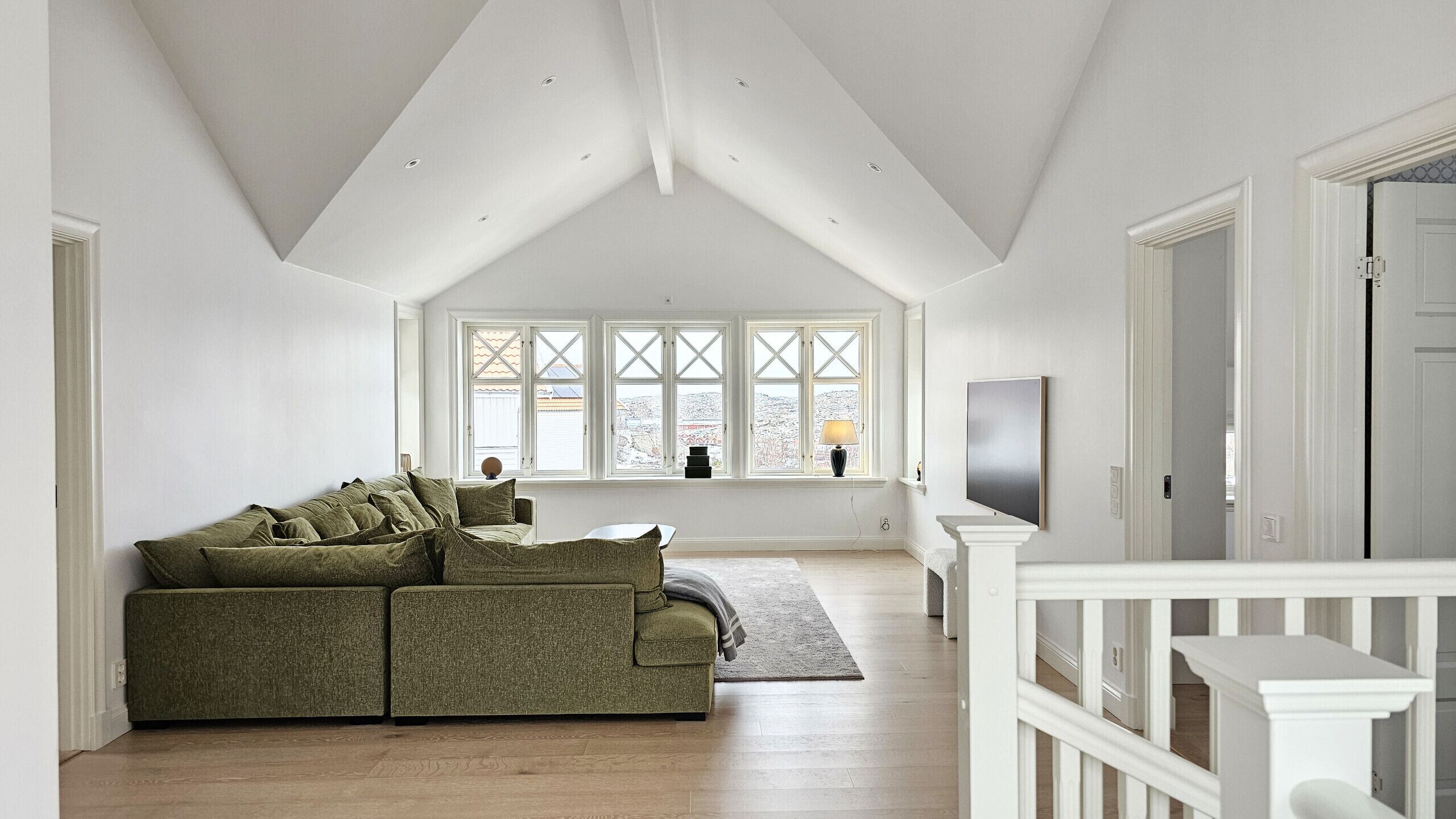 Um amplo loft com pé direito alto, uma janela grande com ventilação cruzada, um sofá verde e uma TV fixada na parede.