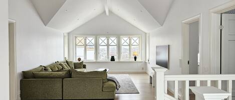 Um amplo loft com pé direito alto, uma janela grande com ventilação cruzada, um sofá verde e uma TV fixada na parede.