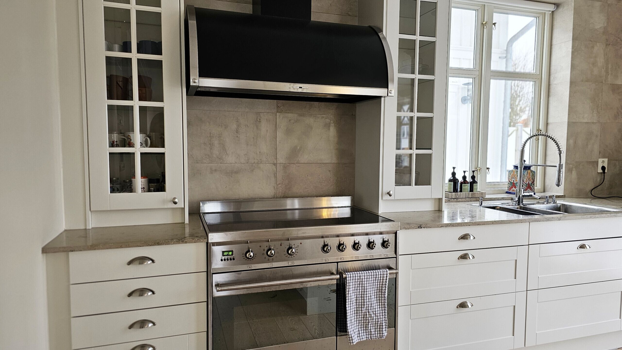 A kitchen with a stainless steel stove, oven, and a black hood. There are white cabinets with glass doors, a window with a view, and a sink with a faucet.
