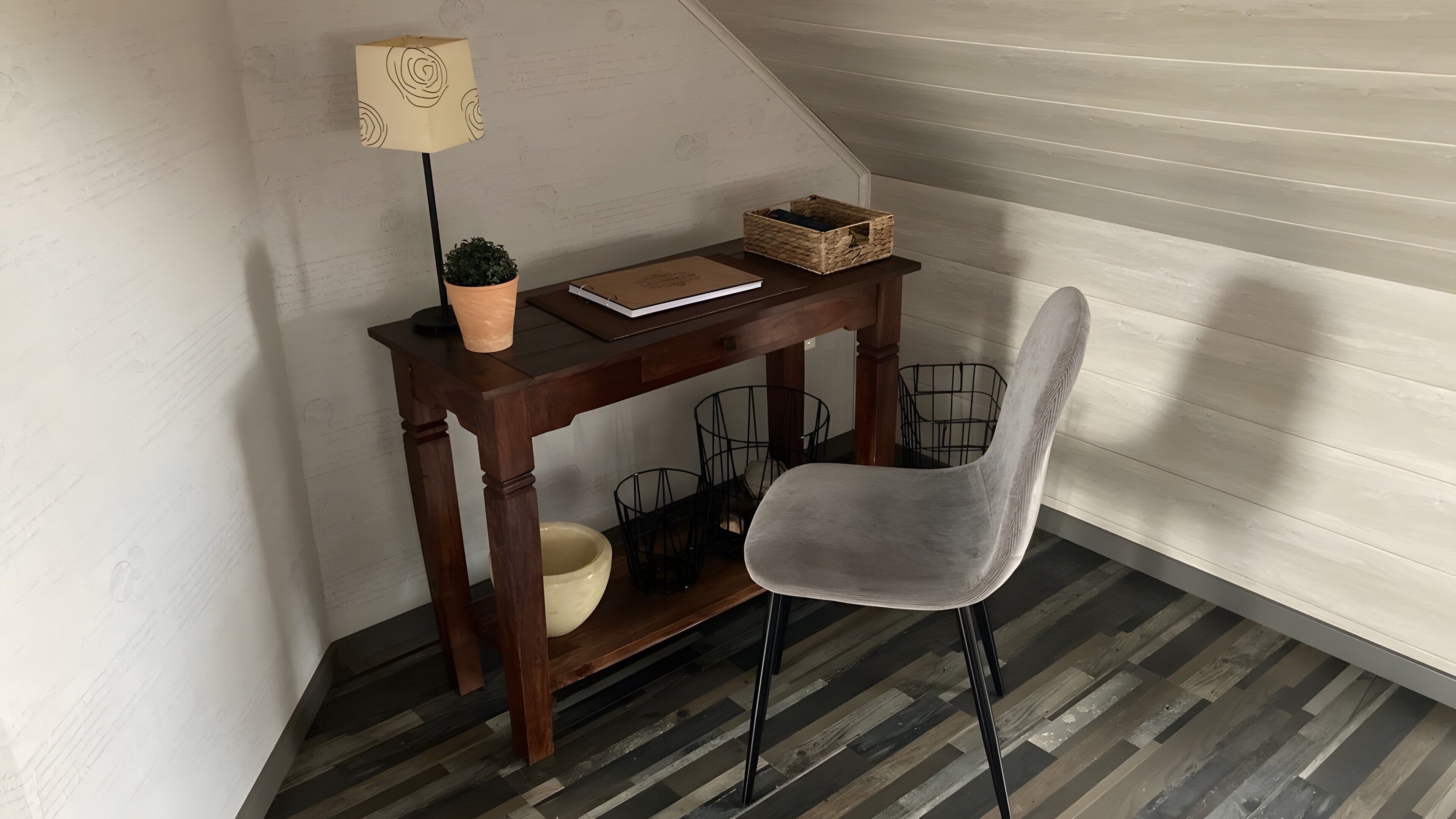 A corner room with a wooden desk, a chair, a lamp, a potted plant, and a basket.