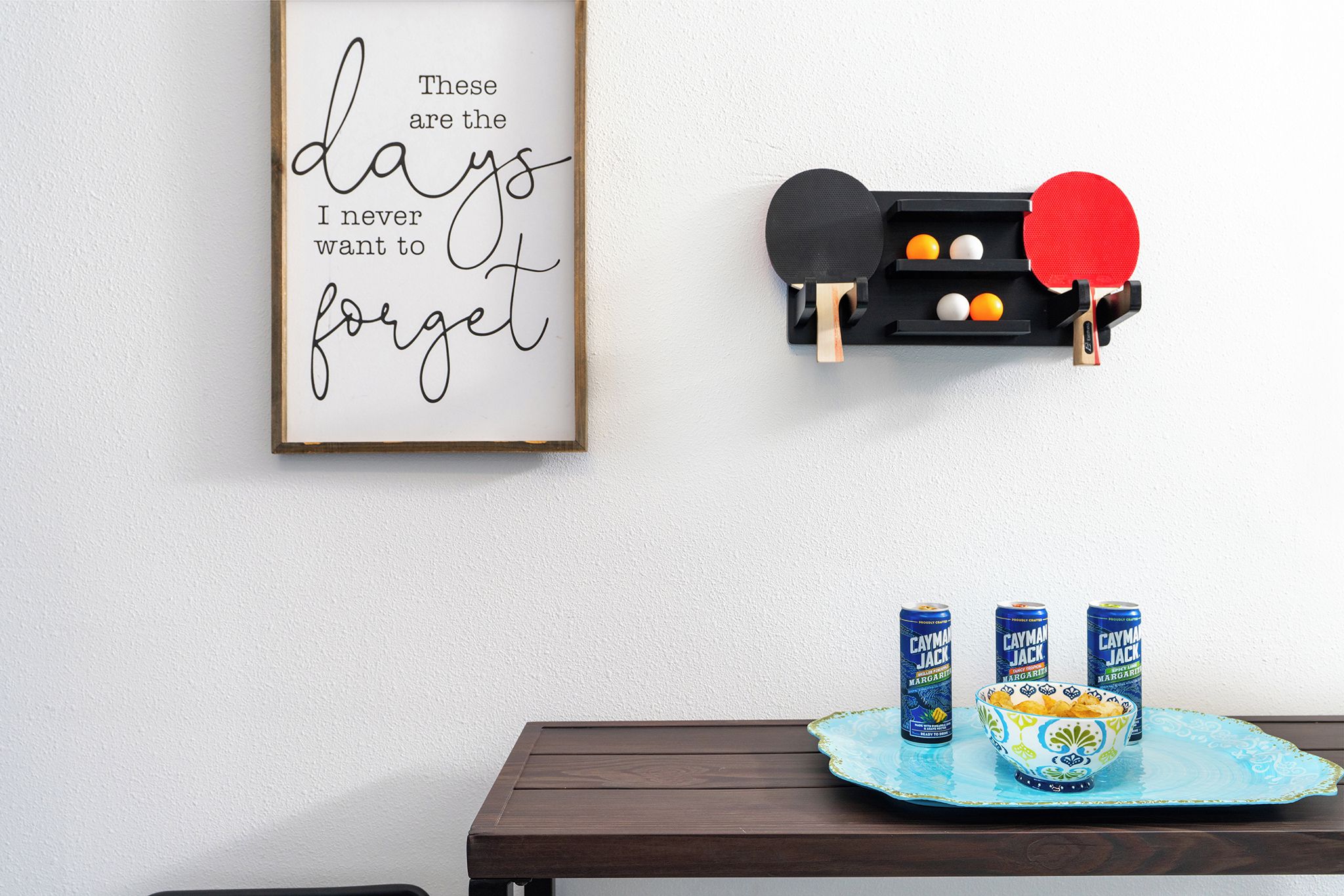 A framed quote on a wall, a ping pong table with paddles, and three cans of Cayman Jack Margarita on a table.