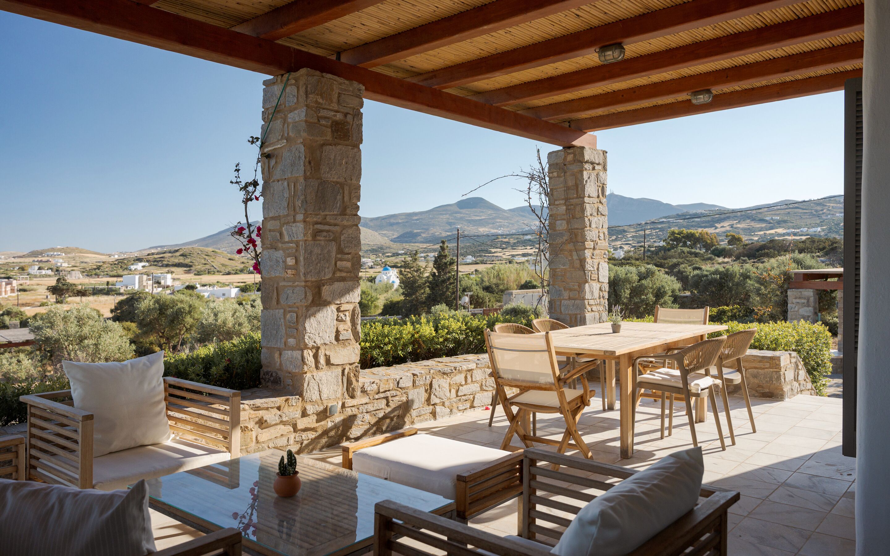 Zona d'estar a l'aire lliure coberta, amb pilars de pedra, una taula i cadires, amb vista a un paisatge idíl·lic amb turons i edificis.