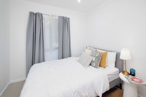 Una cama bien hecha con ropa de cama blanca, una mesita de noche con lámpara y una ventana con cortinas grises.