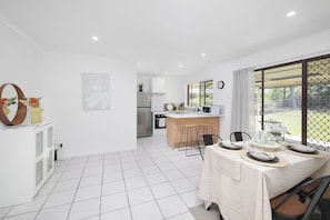 A modern kitchen with white cabinets, a central island, and a dining area with a table set for two. - Furnished 3BR House | Huge Backyard | Weekly Stay Available (Hillcrest)