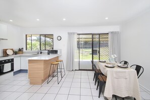 A modern kitchen with a central island, bar stools, a dining table set for four, and a view through large sliding doors. - Furnished 3BR House | Huge Backyard | Weekly Stay Available (Hillcrest)