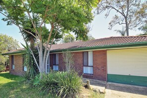 A two-story brick house with a green garage door, surrounded by lush greenery and trees. - Furnished 3BR House | Huge Backyard | Weekly Stay Available (Hillcrest)