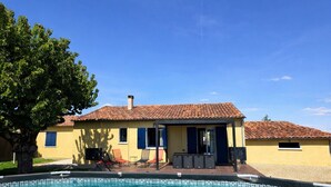A clear swimming pool with a tiled deck, a yellow building with a tiled roof, and a tree in the foreground.
