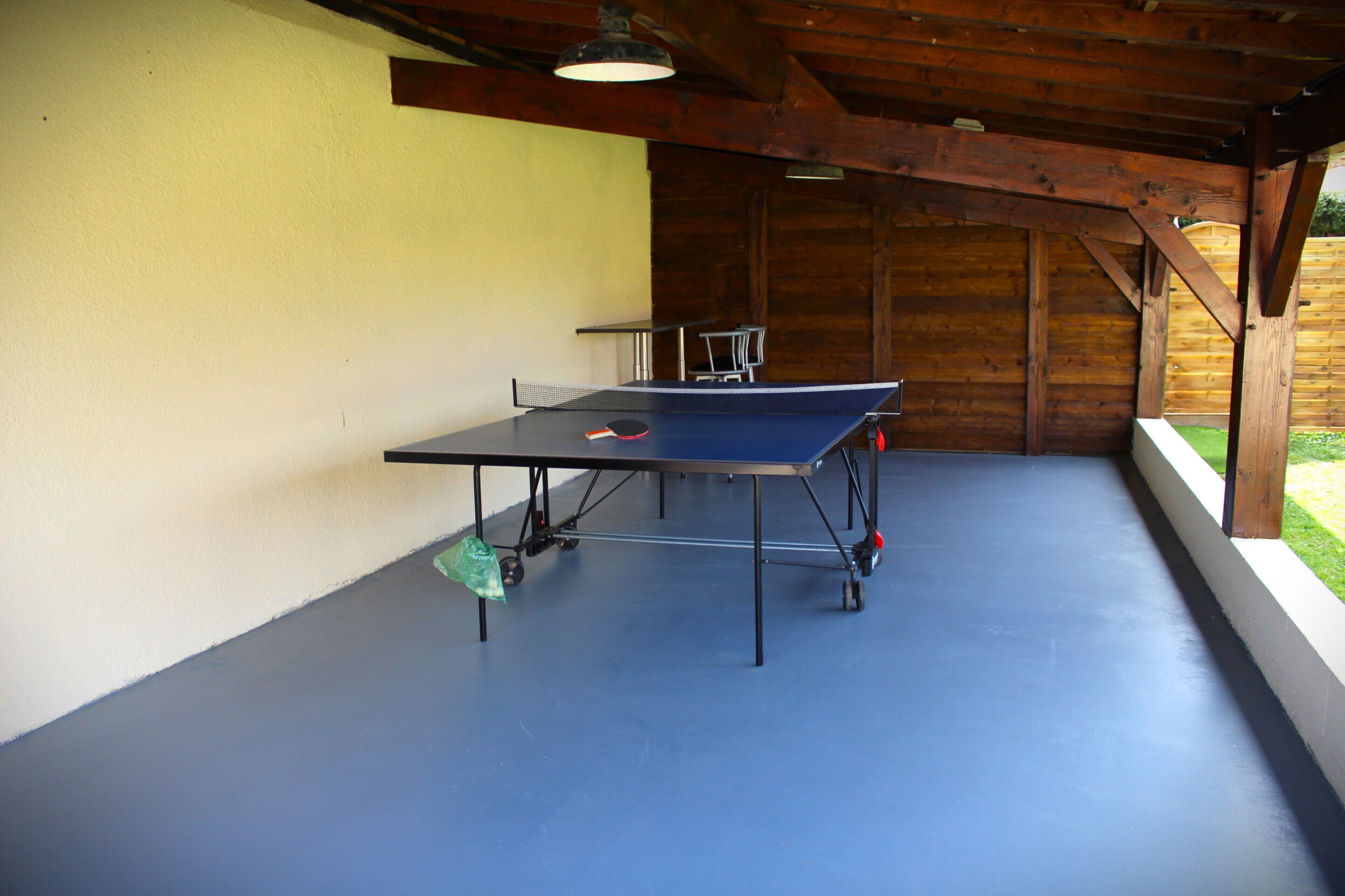 An indoor ping pong table with a blue surface, a net, and a paddle, set against a yellow wall and wooden ceiling.