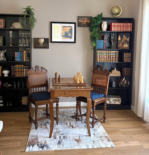 A room with a chess table, two chairs, a bookshelf, and a framed picture on the wall.