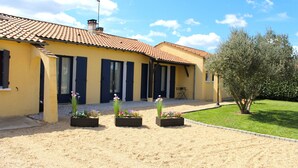 A yellow building with a tiled roof, dark shutters, and a garden with flower beds.