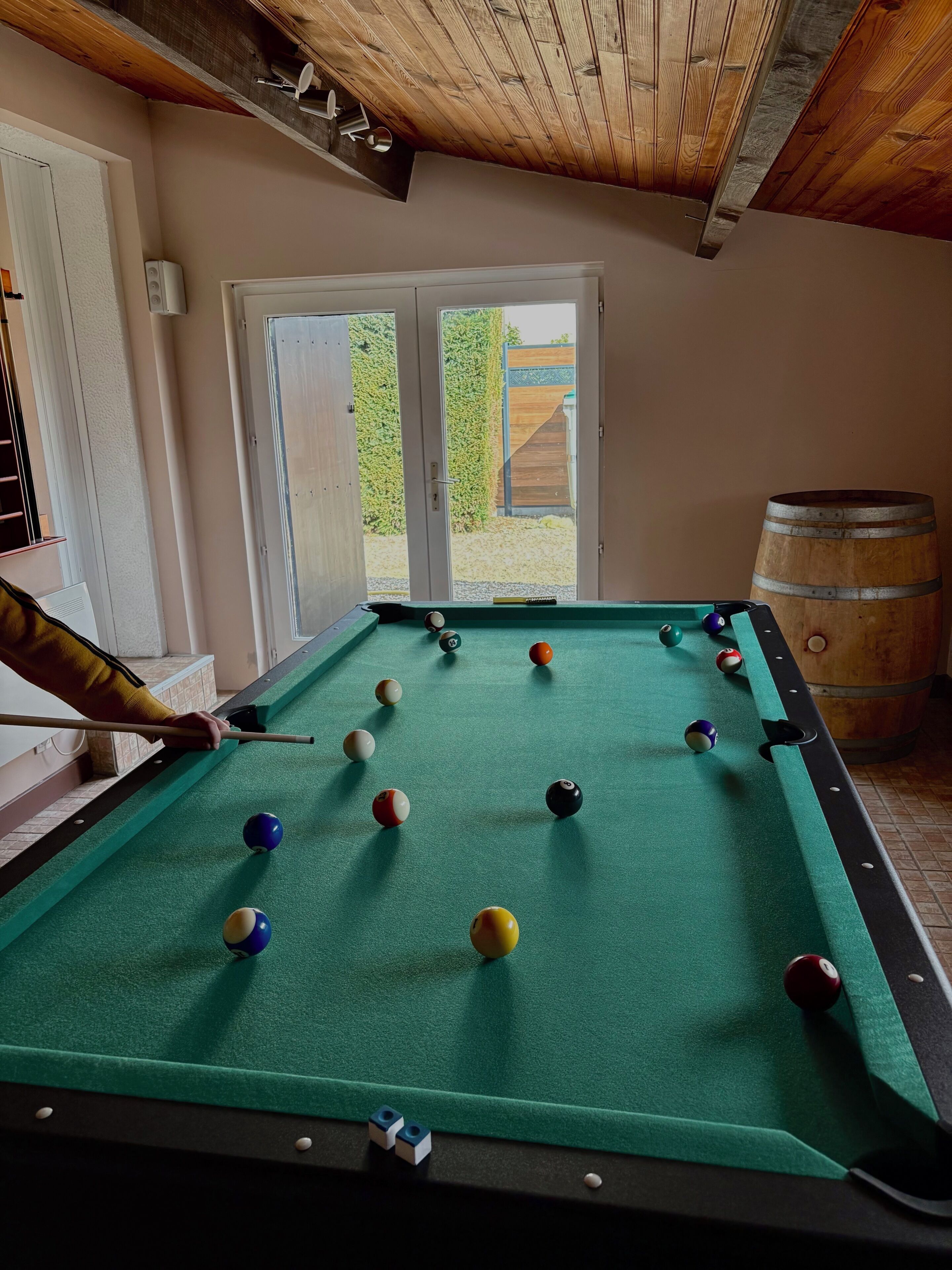 Une table de billard avec des billes, un tonneau en bois et une porte donnant sur l’extérieur.