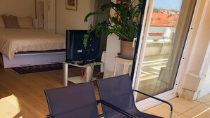 Ein Zimmer mit einem Bett, einem Schreibtisch, einem Stuhl, einer Topfpflanze und Blick durch eine Glastür.
