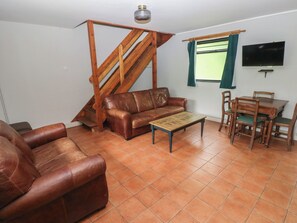 Una sala de estar con un sofá de cuero marrón, una mesa de centro de madera, una mesa de comedor con sillas, una escalera de madera y un televisor montado en la pared.