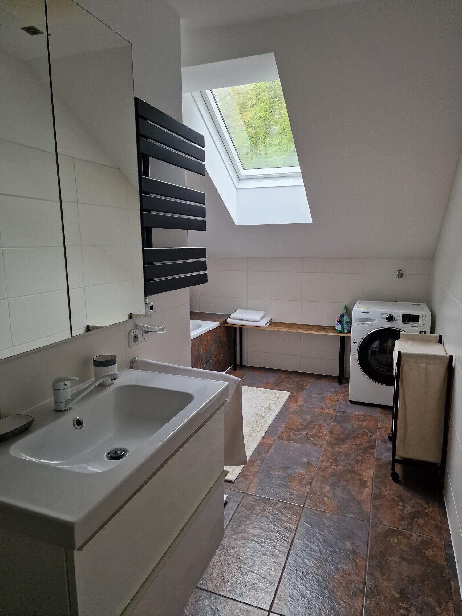A compact, modern bathroom with a large mirror, a bathtub, a washing machine, and a skylight.