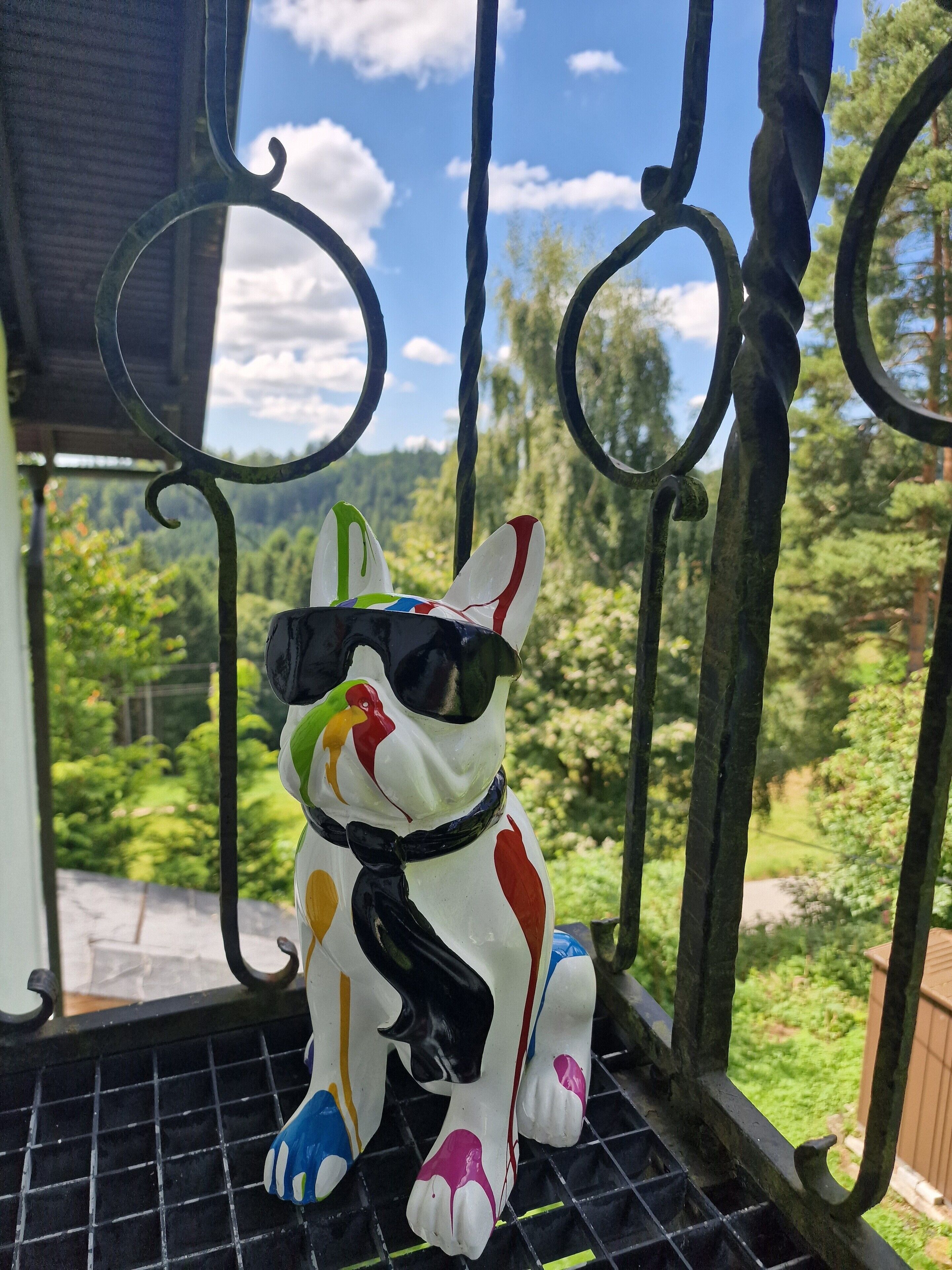 Une figurine de chien aux couleurs vives, posée sur la rambarde d’un balcon, avec vue sur un paysage verdoyant et un ciel dégagé.
