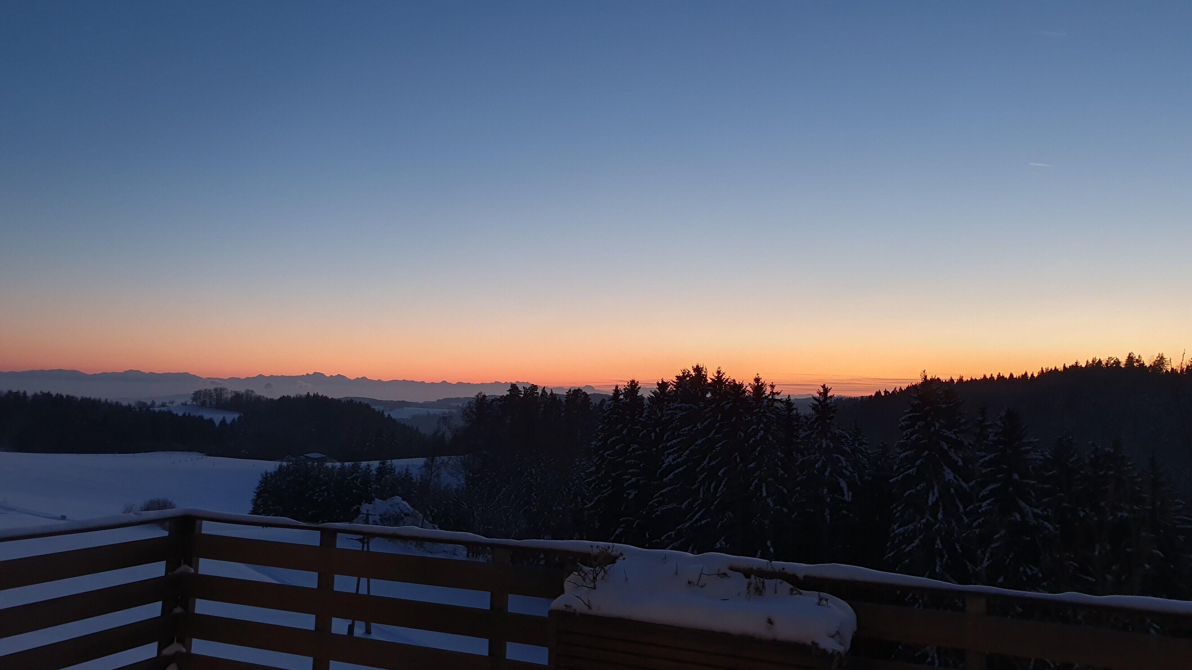 Un paysage enneigé avec une clôture en bois au premier plan et un coucher de soleil en arrière-plan.